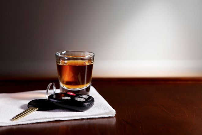 Shot glass of amber liquid, car keys, and white napkin on a wooden table, against a gray and white background.