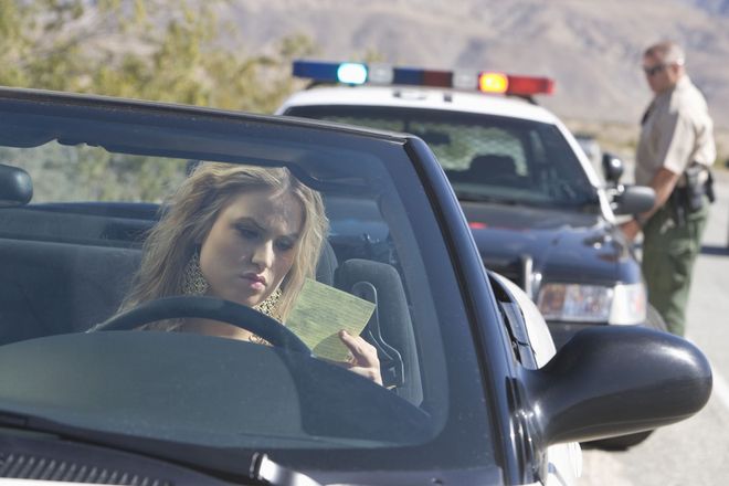 Woman in car looks at a document; police officer stands by patrol car on roadside.
