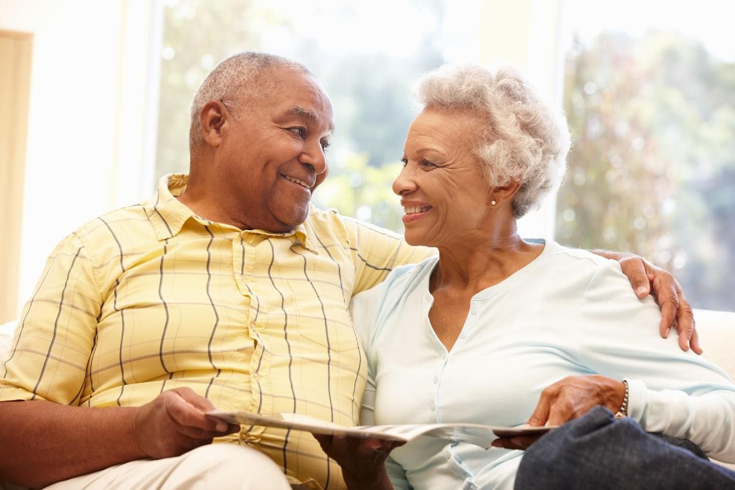 Smiling couple on a sofa; man with arm around woman, looking at a magazine. Natural light.