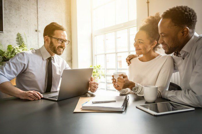 Advisor showing laptop to a couple at table; sunny office setting.