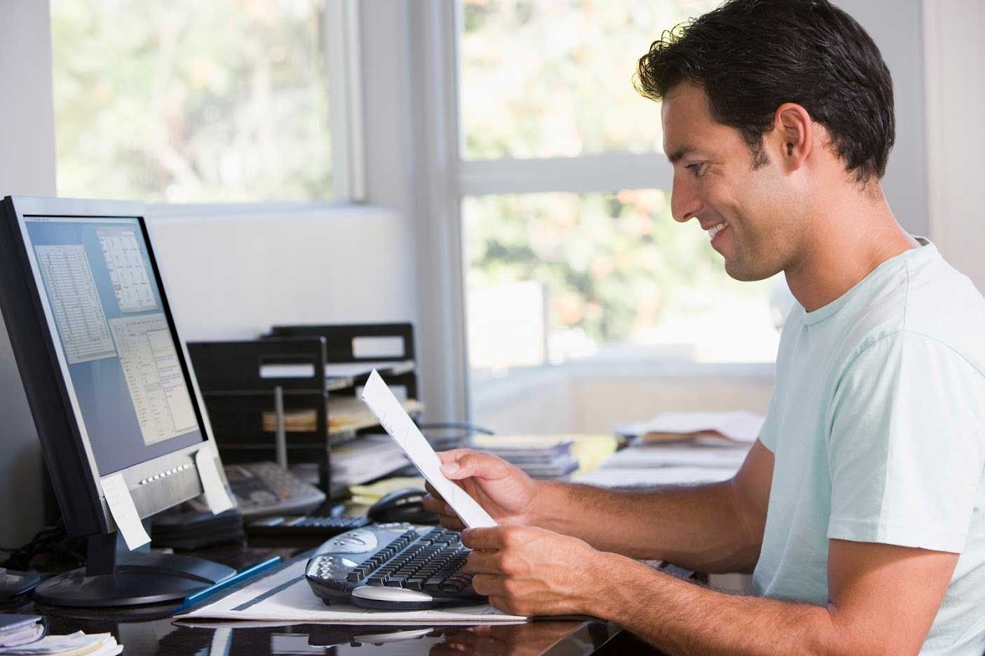 Man smiling while looking at paper, sitting at a desk with computer, near a window.