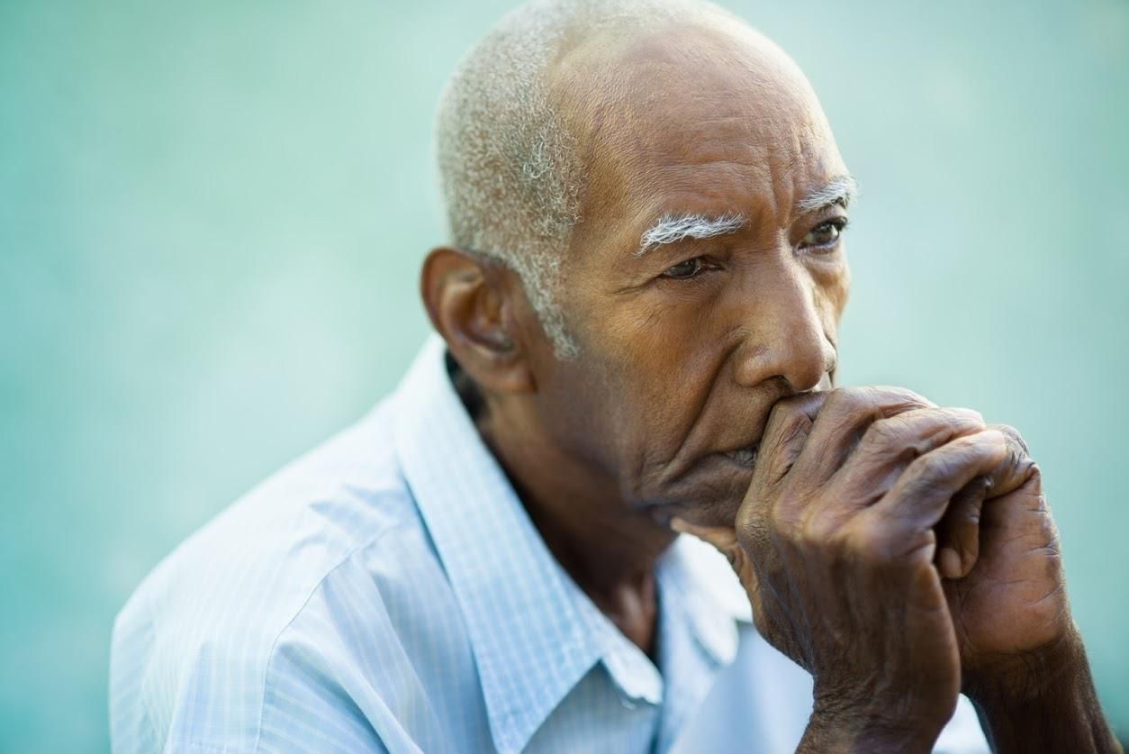 Older person with hands near face, looking down, contemplative expression.