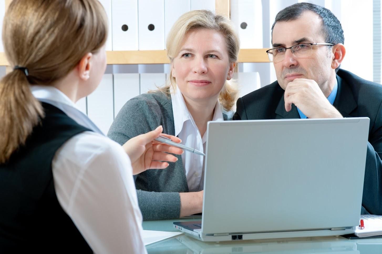 A professional woman gestures with a pen, presenting information on a laptop to a couple at a desk.