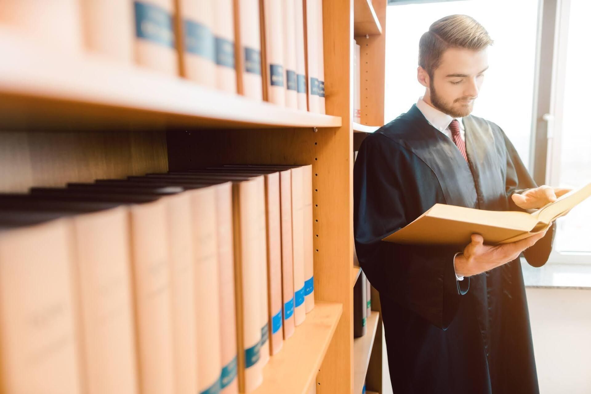 Lawyer in black robe reads a book in a library, lit by sunlight. Bookshelves in the foreground.