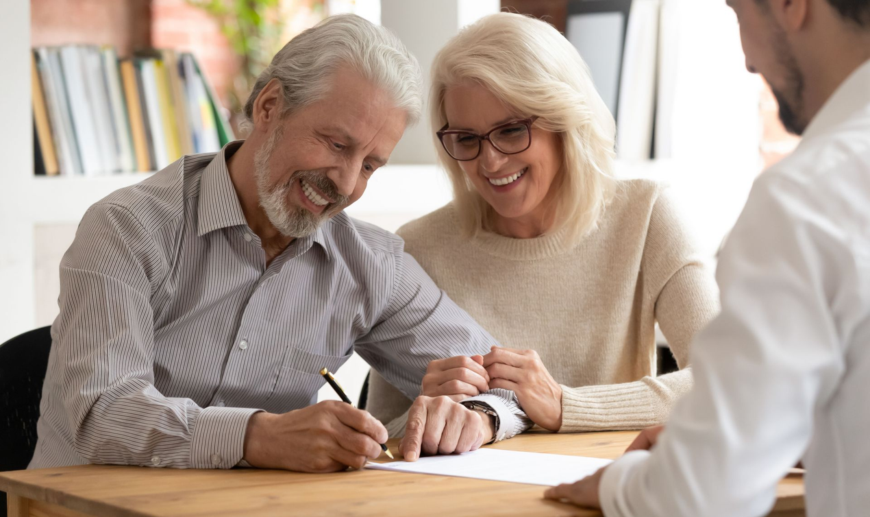Couple signing documents with an advisor at a desk; smiling, indoors.