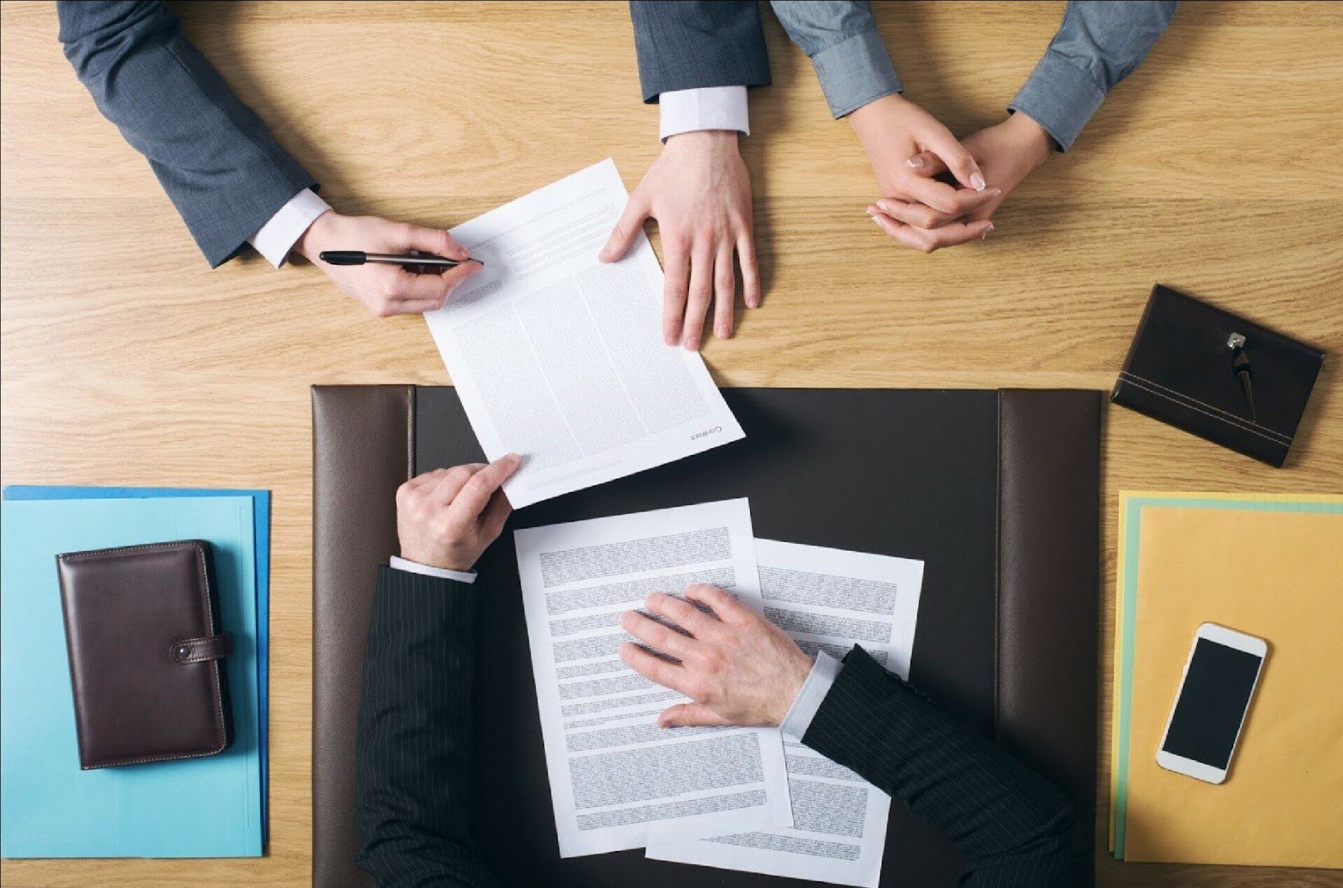 Hands signing a document, with others present at a wooden desk with papers, notebooks, and a phone.
