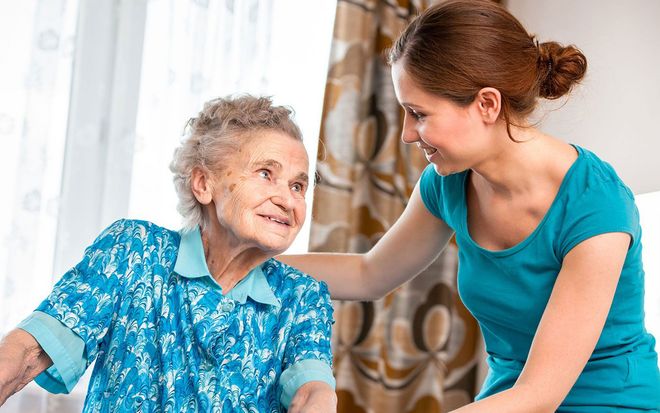 Caregiver in teal shirt gently holds an elderly woman’s shoulder indoors near a window.