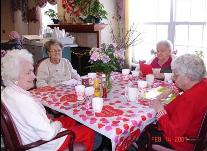 Five elderly women sit around a table with flowers and drinks in a bright dining room.