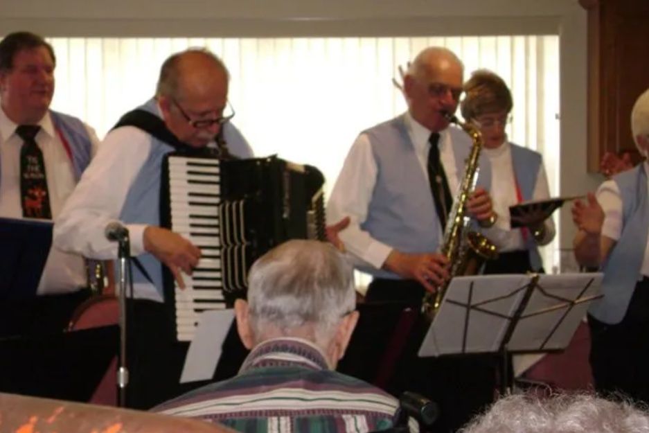 Small band performing indoors, with accordion, brass instruments, and music stands before an audience