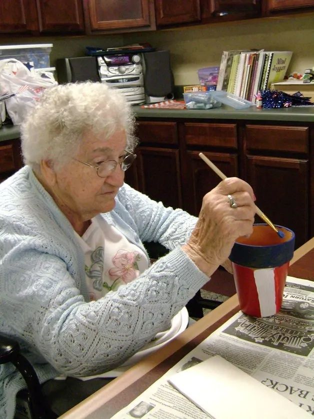 Elderly woman painting a red-and-blue cup at a table in a kitchen.