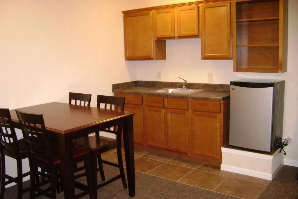 Small kitchen with wooden cabinets, sink, fridge, and a dining table with chairs on tiled floor