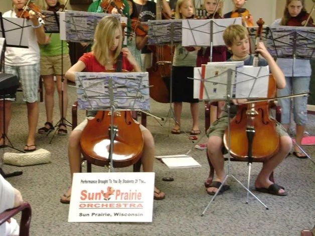 Children seated with cellos and music stands during an outdoor orchestra rehearsal