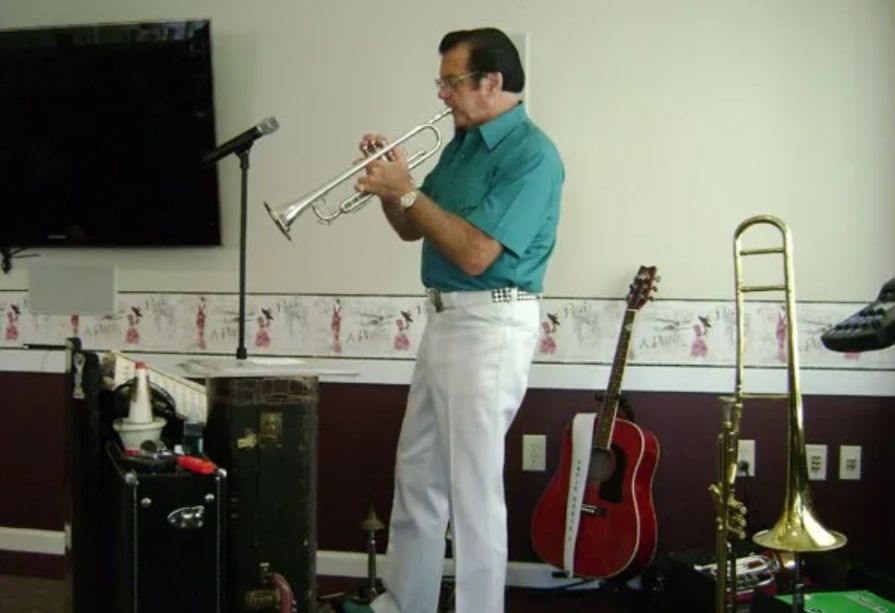 Man playing trumpet on a small stage with a TV, guitar, and trombone in the background