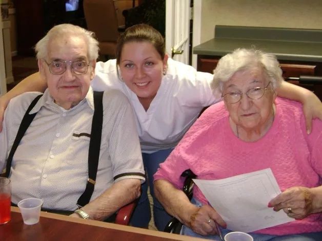Three people seated at a table, smiling, with a woman in pink holding papers and a young woman standing behind them.