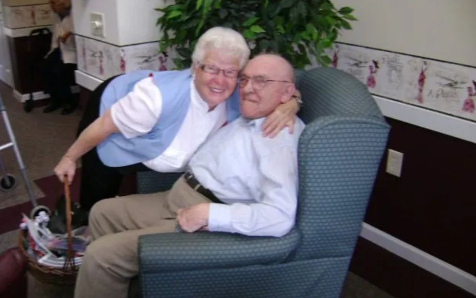 Two smiling people seated together in a patterned armchair, embracing in a cozy room.