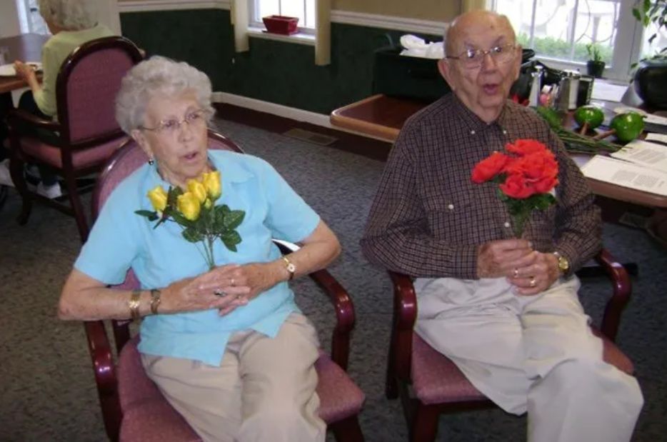 Two elderly people seated indoors, each holding a bouquet of flowers.