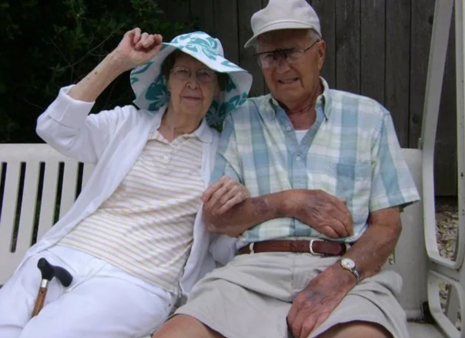 Two people sitting on a white bench, one adjusting a blue hat, outdoors by a wooden wall