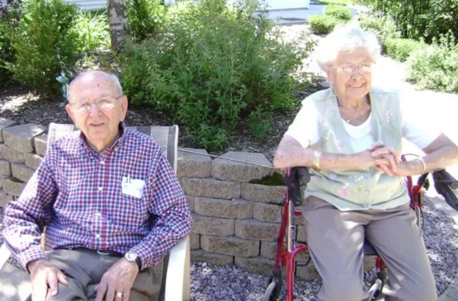 Two older adults sitting outdoors in chairs beside a stone wall and shrubs, smiling in the sun