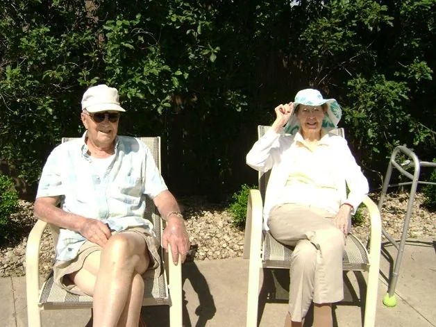Two people sitting outdoors on lawn chairs, one adjusting a hat in bright sunlight.