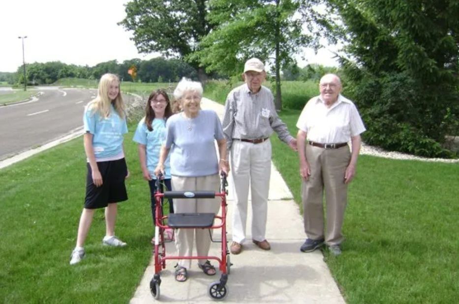 Five people standing on a sidewalk outdoors near a road and green lawn; one uses a walker.