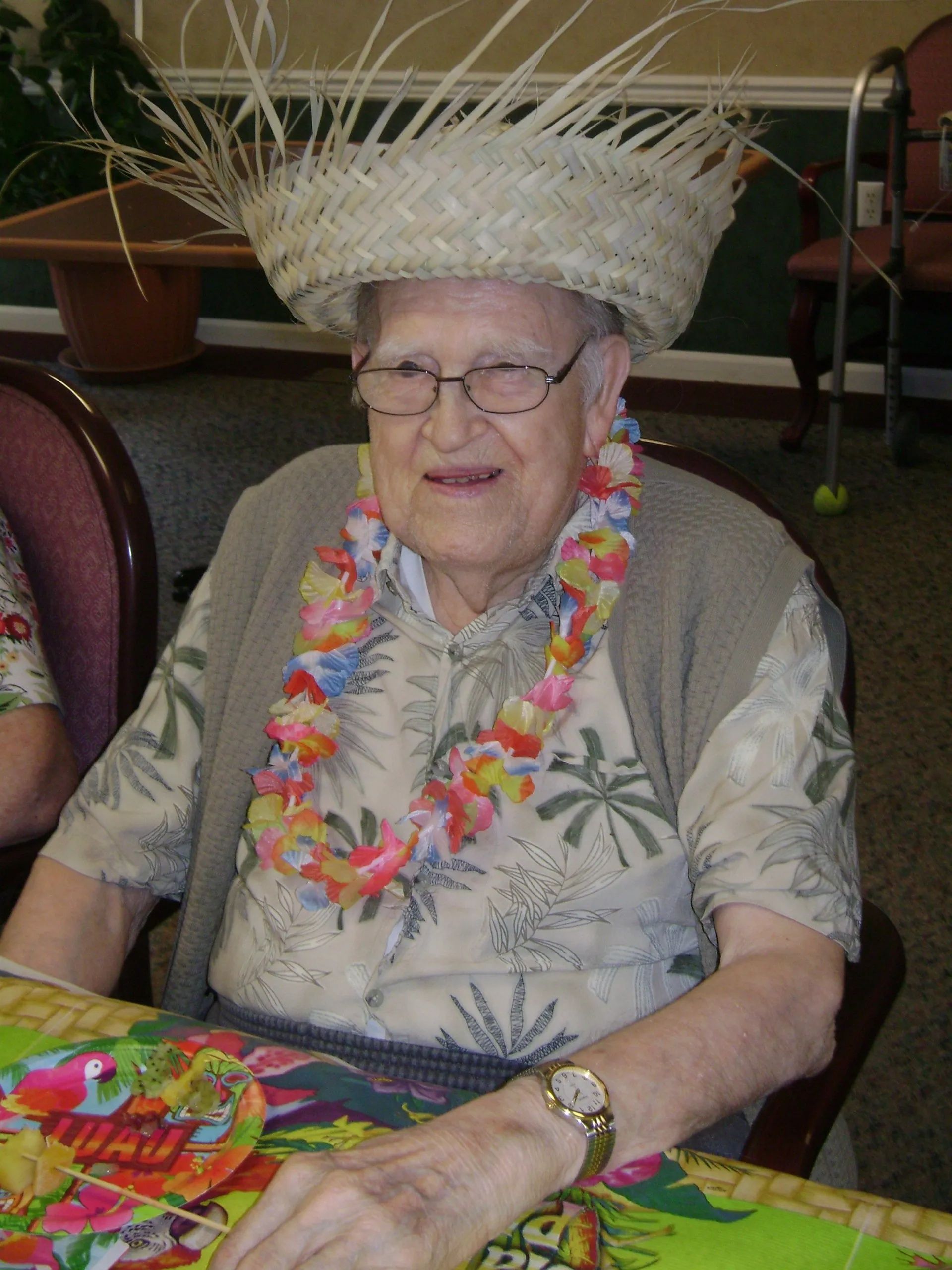 Smiling elderly woman wearing a straw hat and colorful leis, seated at a table in a decorated room