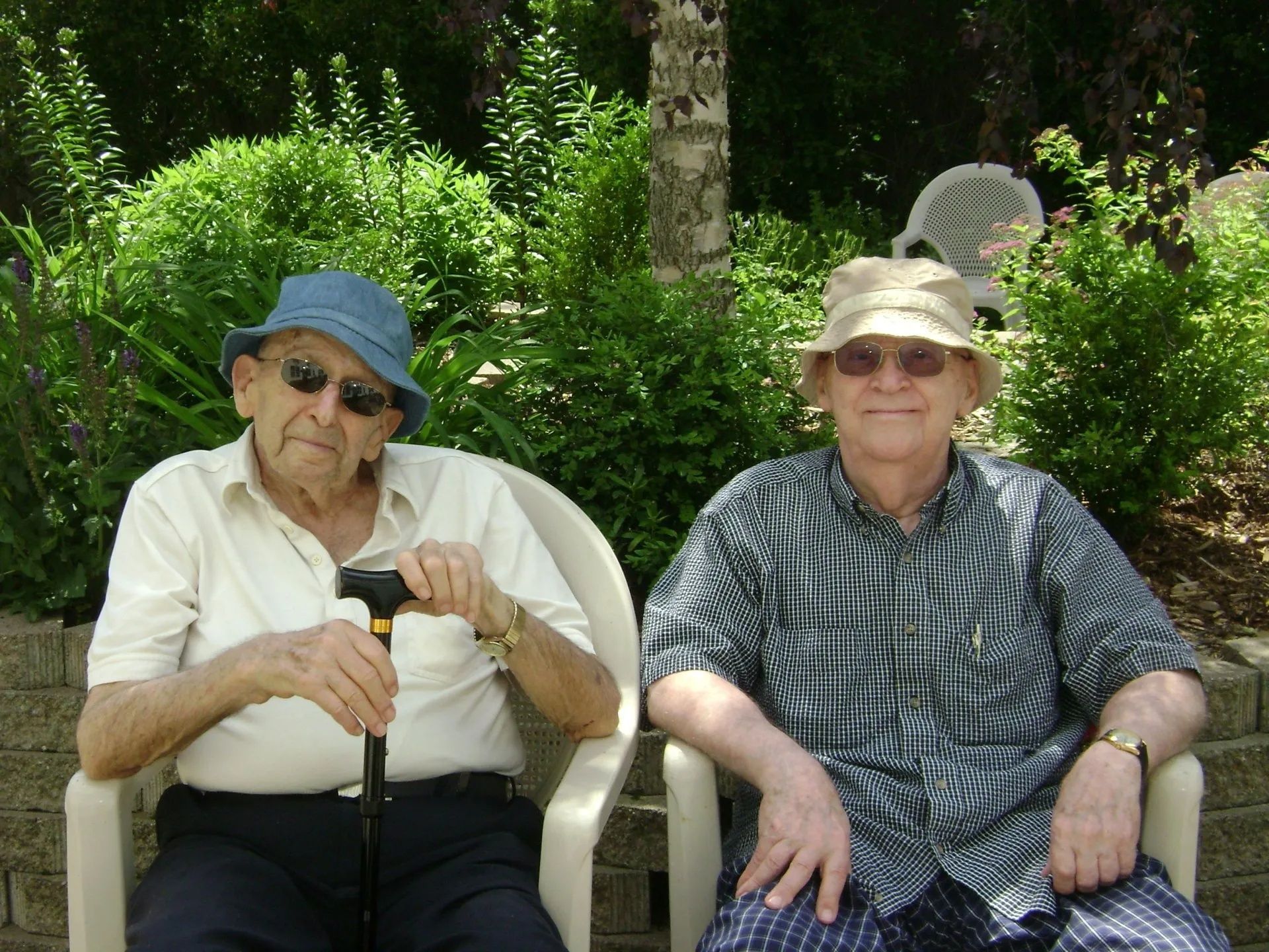 Two people sitting in lawn chairs outdoors in a leafy garden, both wearing hats and sunglasses.
