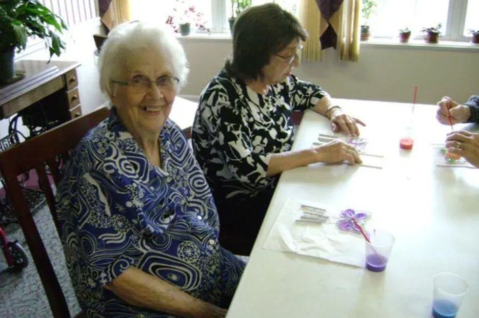 Two women sitting at a table in a bright room, one smiling, with craft supplies nearby