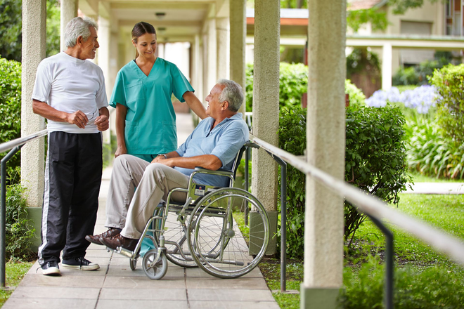 Caregiver in teal scrubs speaking with a seated man in a wheelchair beside an elderly man outdoors on a walkway
