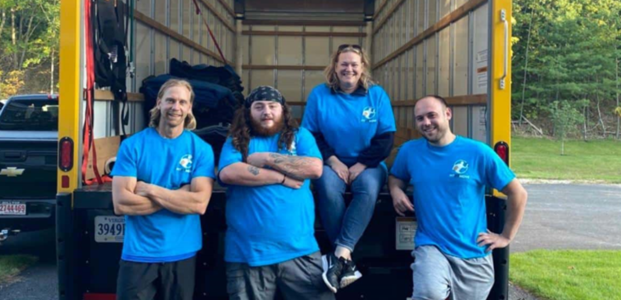 Four movers in blue shirts pose inside a truck; two men crossed arms, one sits and one has hand on hip.