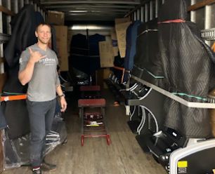 Man in gray shirt gives a shaka sign in a packed moving truck, surrounded by furniture and boxes.