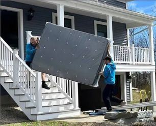 Two people carrying a large mattress down the stairs of a house with a porch.