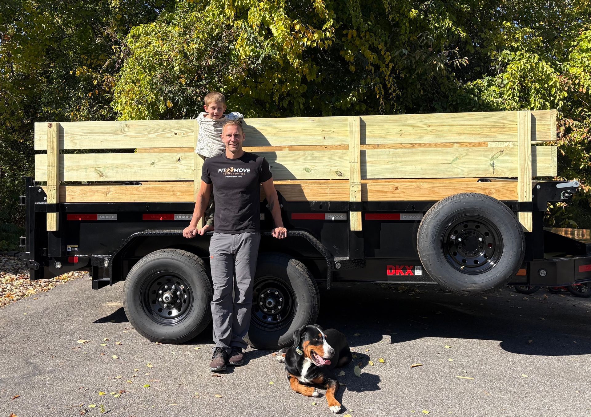 Two people with a dog standing by a trailer loaded with wooden boards outdoors