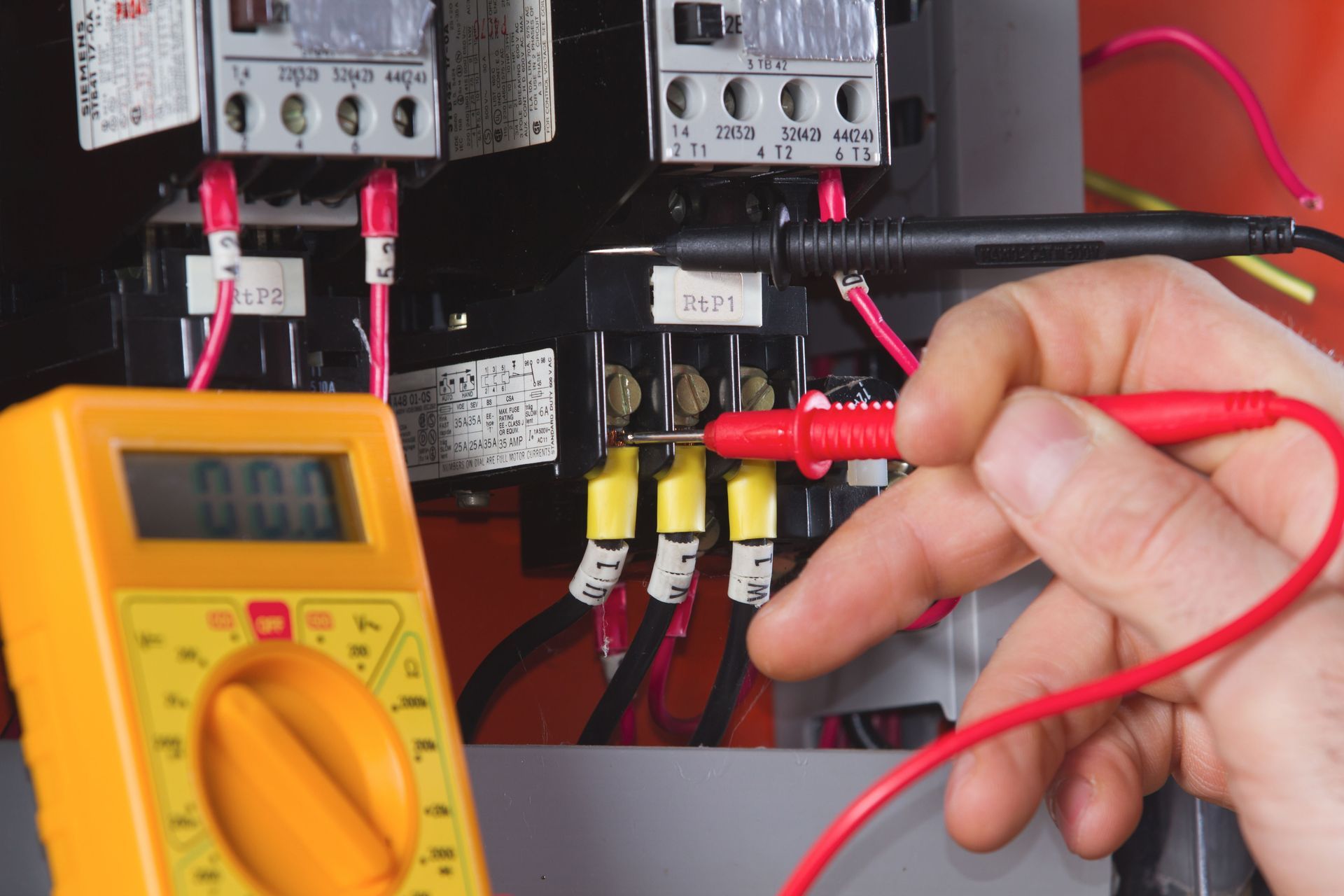 Red and black multimeter probes testing electrical wiring in a panel near switches and relays