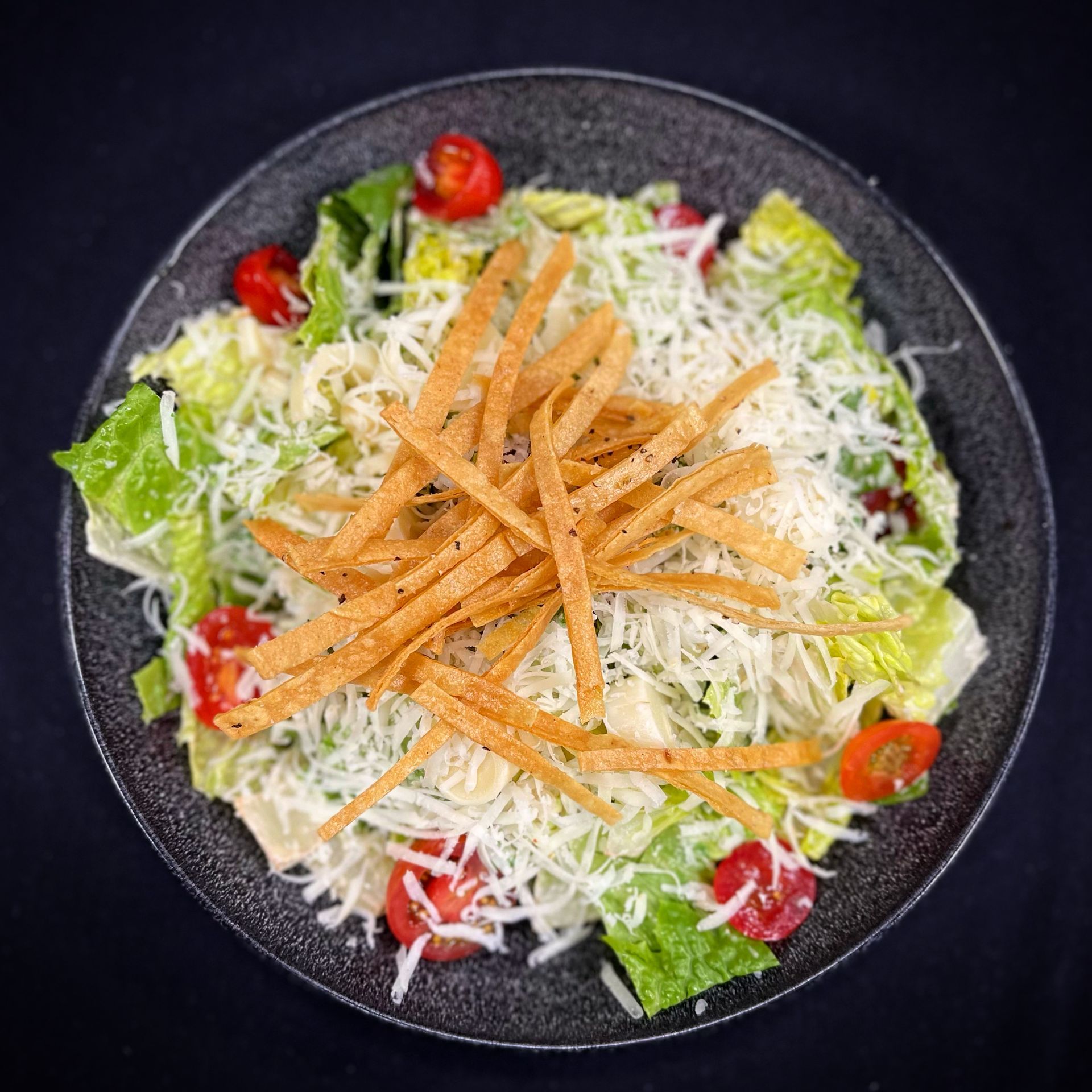 A salad with lettuce , tomatoes , cheese and tortilla strips on a black plate.
