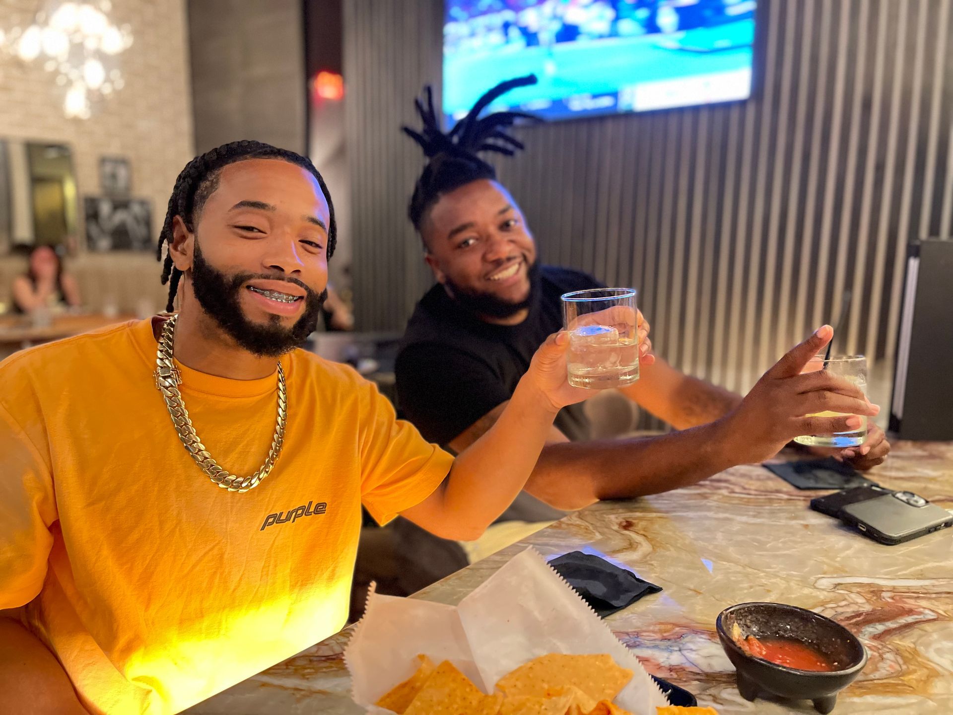 Two men are sitting at a table holding drinks and eating chips.