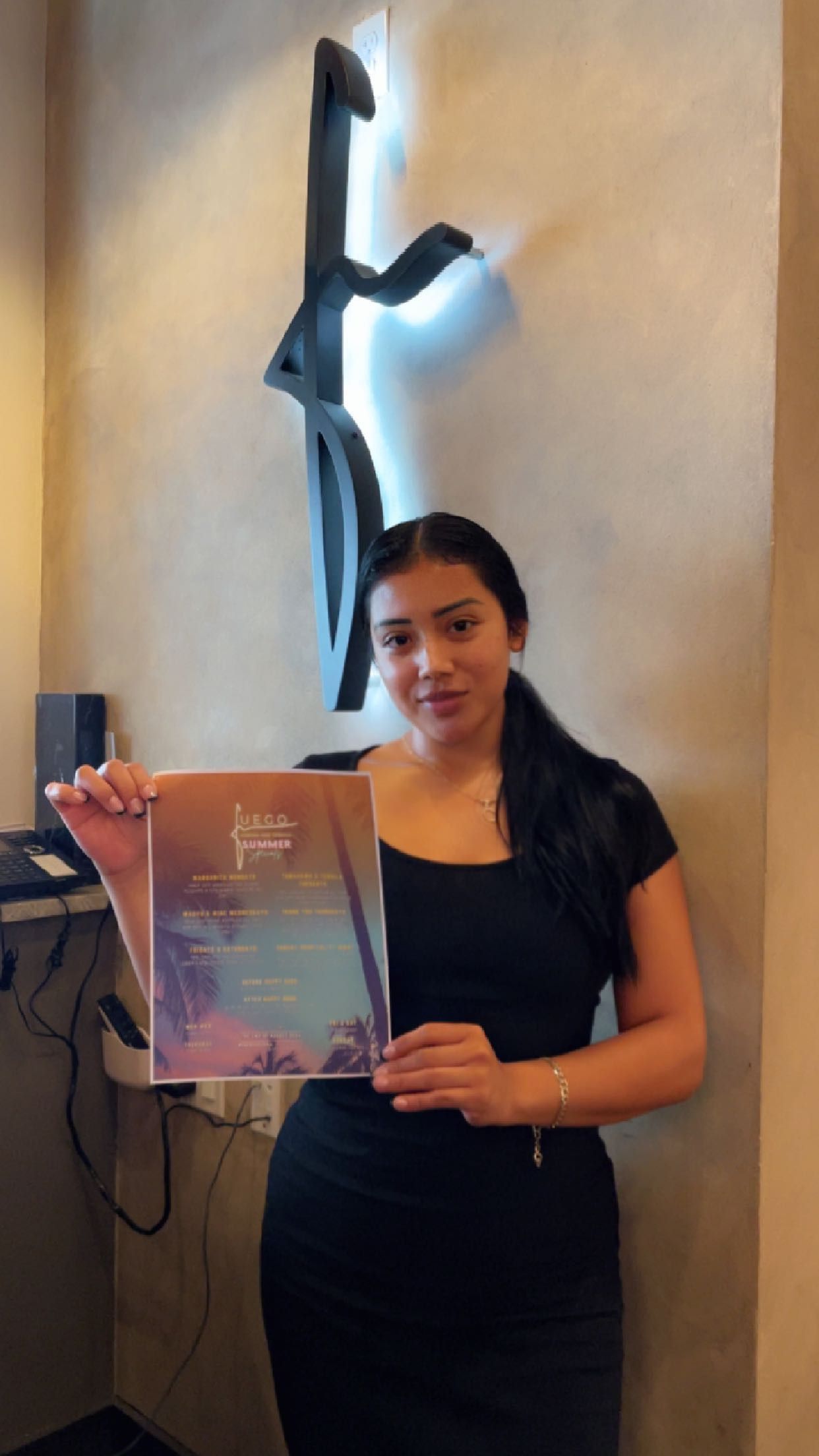 A woman in a black dress is holding a book in front of a wall.