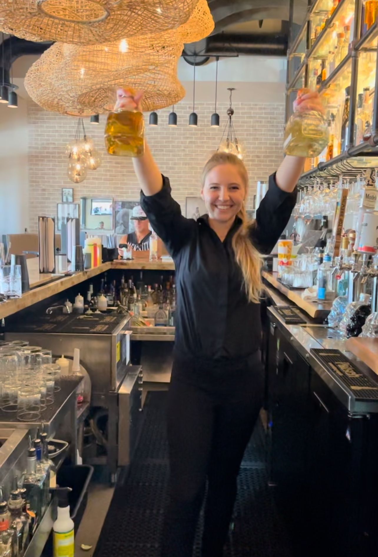 A woman is standing in a bar holding two glasses in her hands.