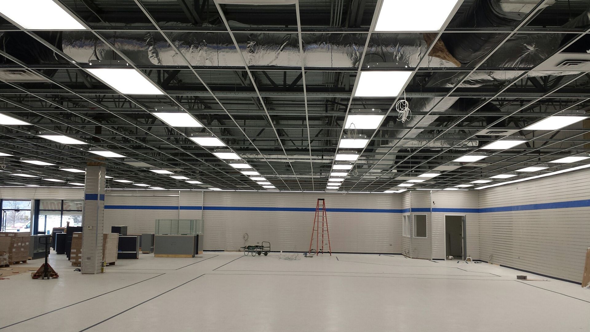 A large, empty commercial space under construction with an exposed grid ceiling, white floor, and a blue stripe on walls.