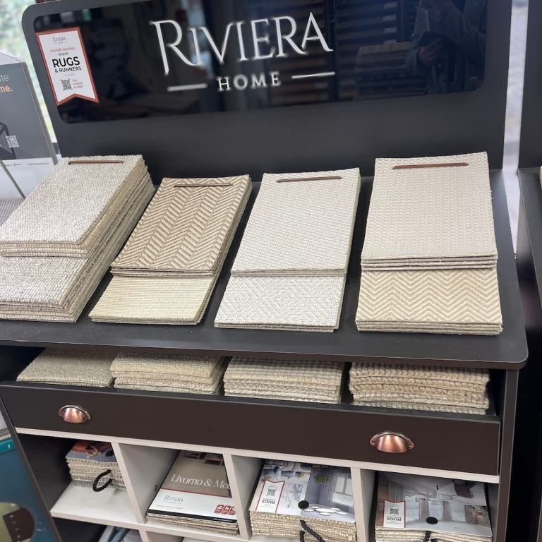 Display of Riviera Home rugs in various neutral tones, stacked on a shelf.