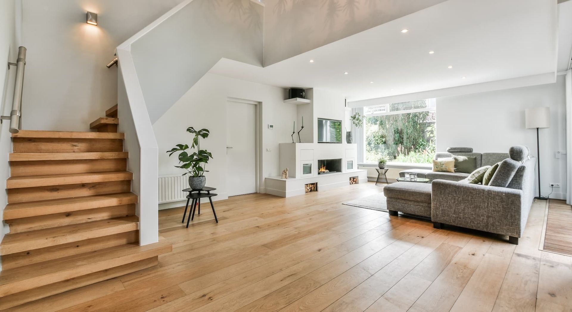Spacious modern living room with wooden floors, stairs, and a gray sofa.