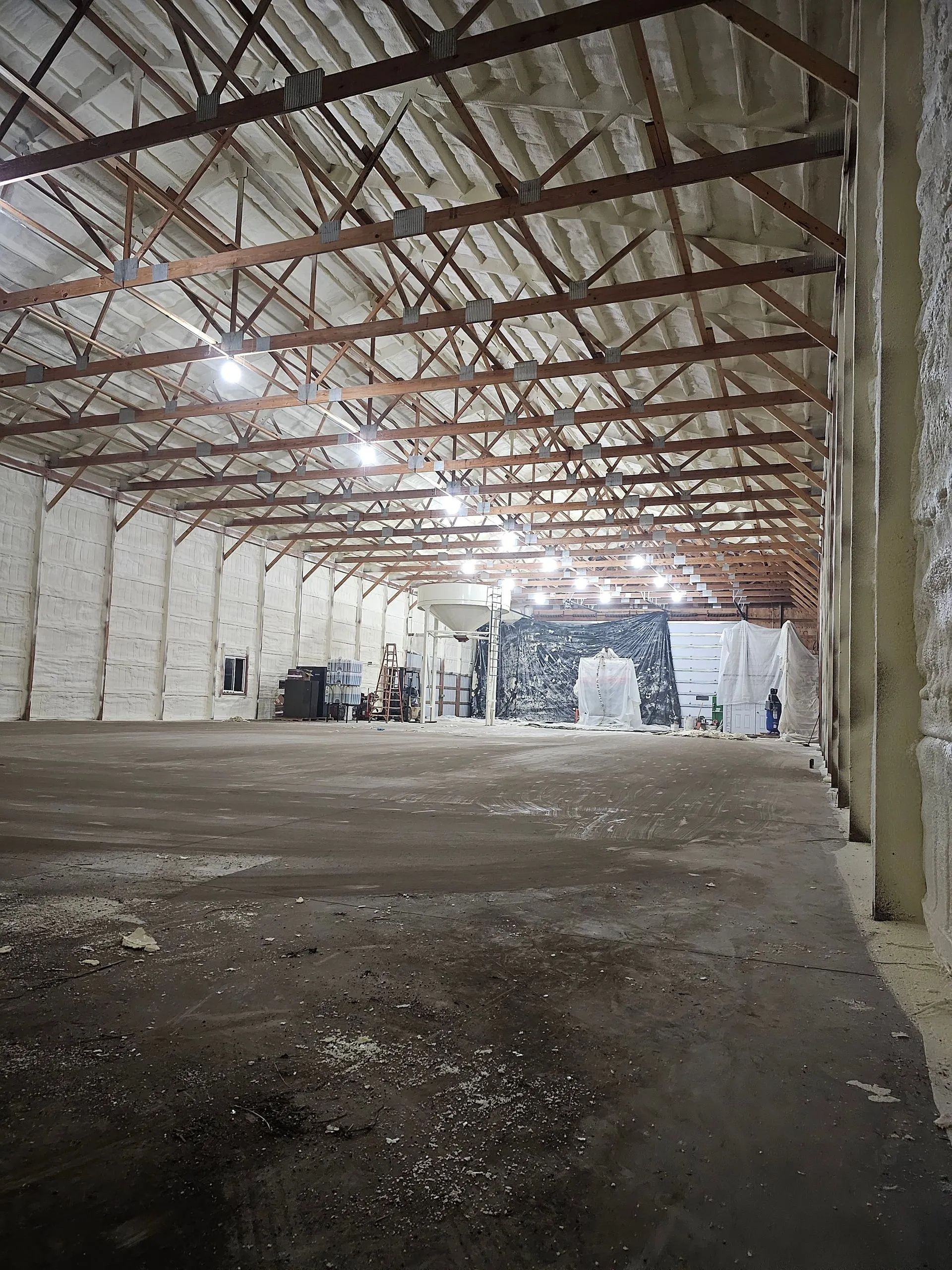 An interior view of a large, empty warehouse with high wooden trusses, spray-insulated walls, and a dirt floor.