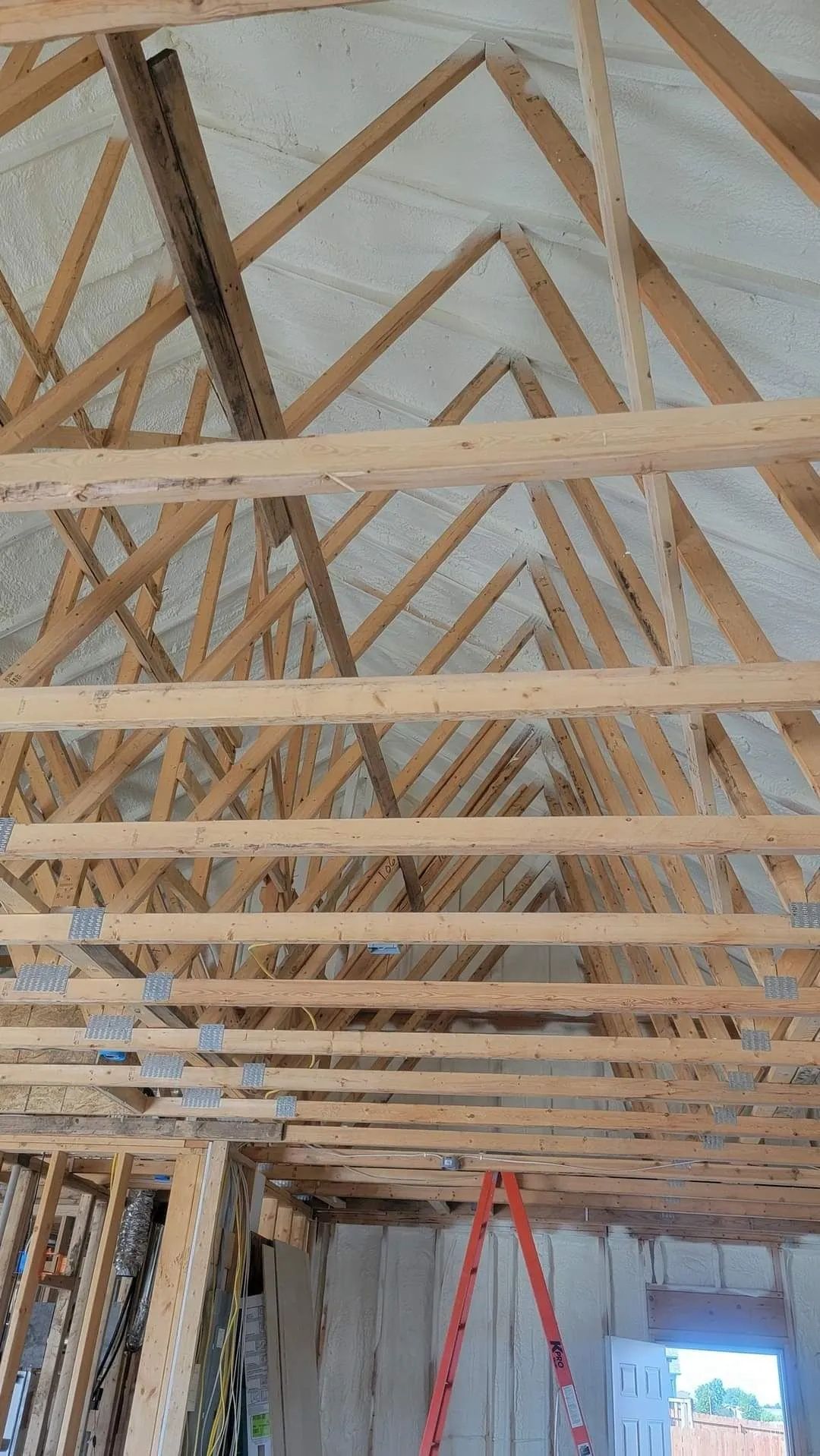 An unfinished attic interior showing wooden roof trusses and white spray foam insulation applied to the roof deck.