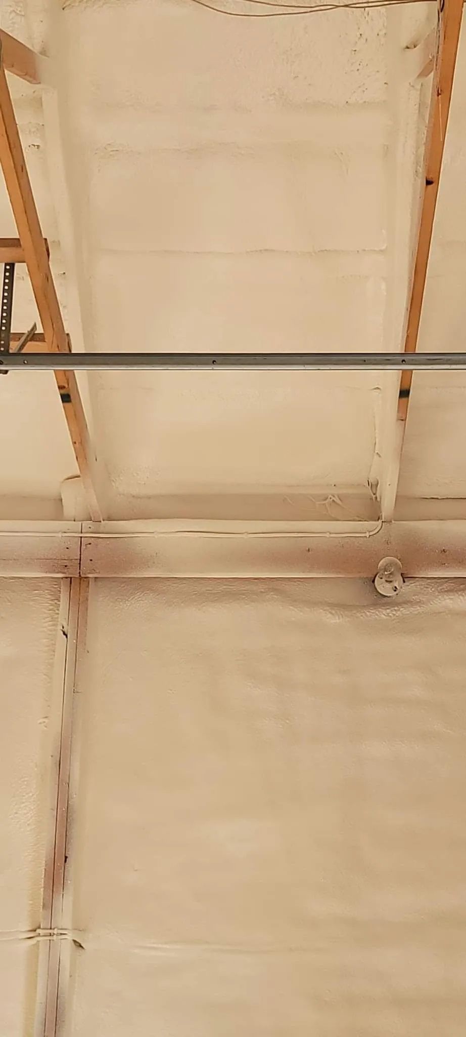 View looking up at a garage ceiling insulated with light-colored spray foam between wooden trusses and a metal bar.