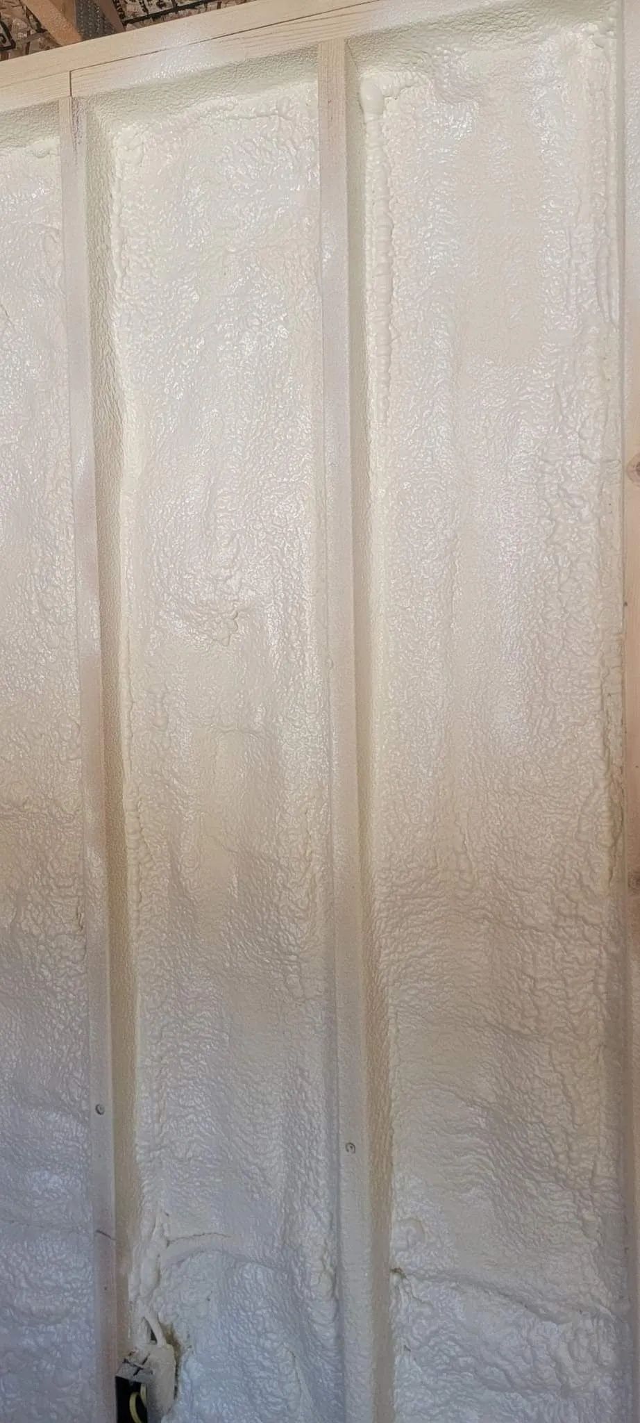Textured, light-colored spray foam insulation installed between vertical wooden wall studs.