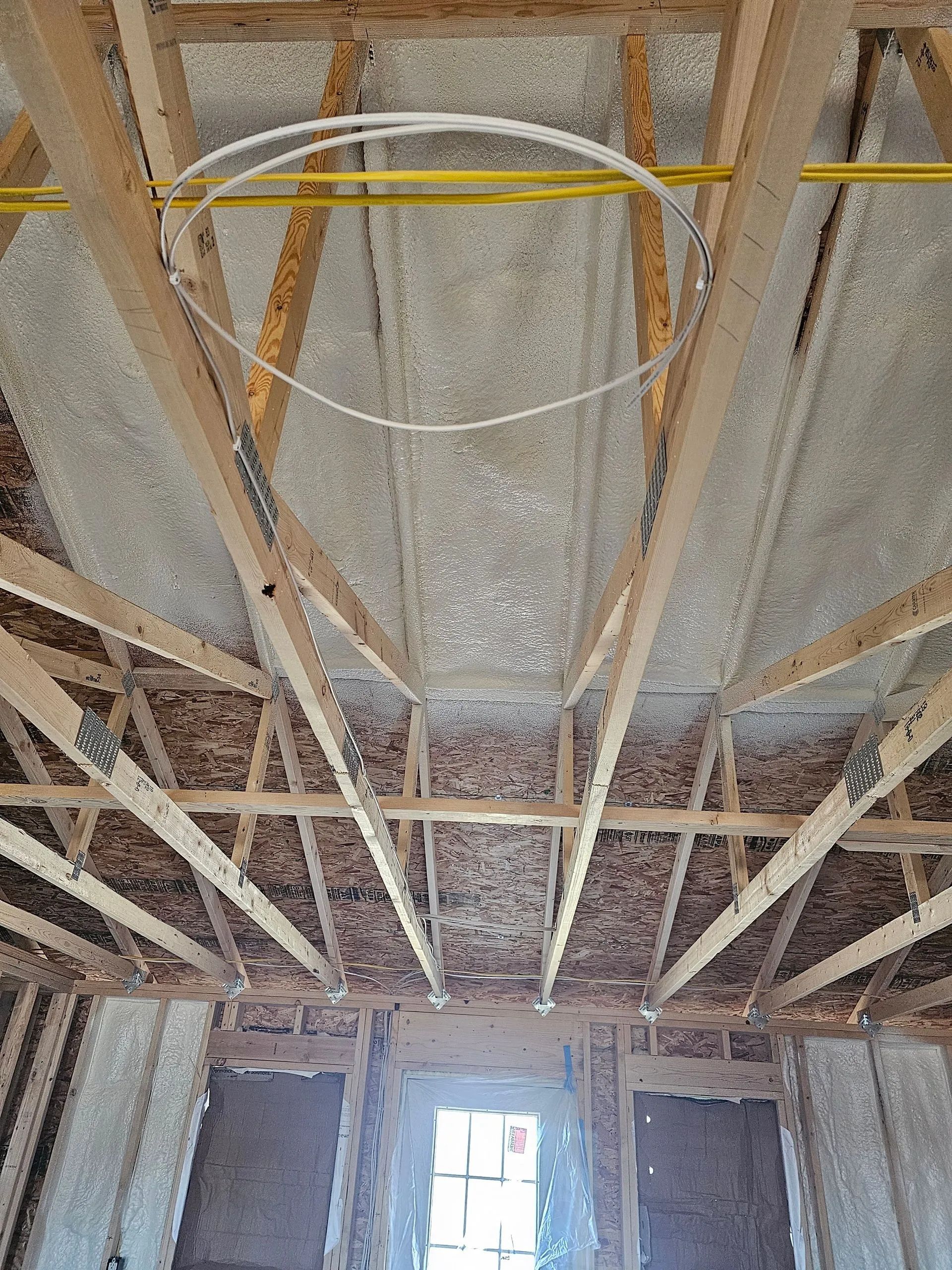 Wooden ceiling trusses and wall frames inside an unfinished building with white spray foam insulation applied.