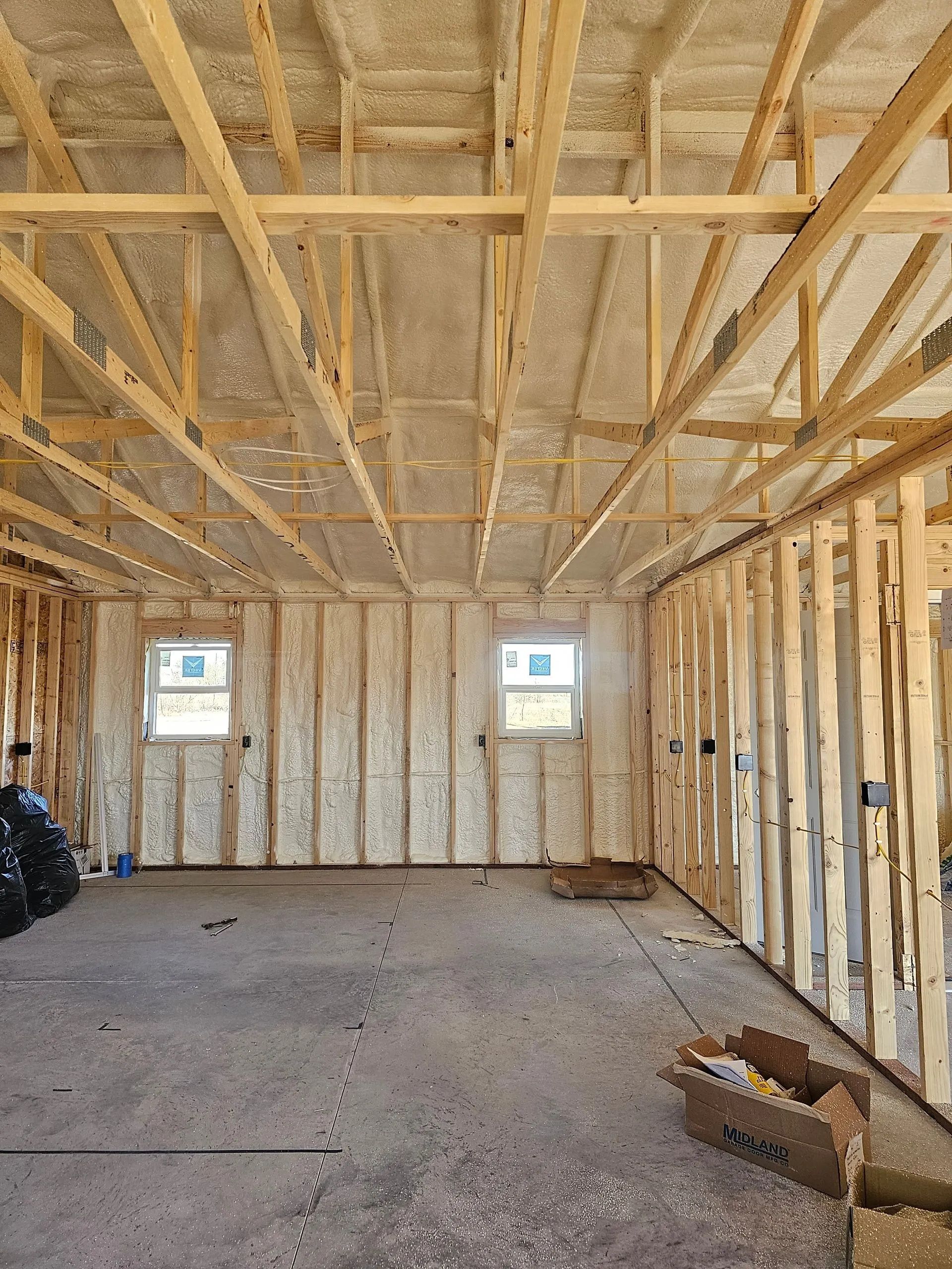 A view inside a building under construction, showing wooden wall studs, roof trusses, and spray foam insulation.