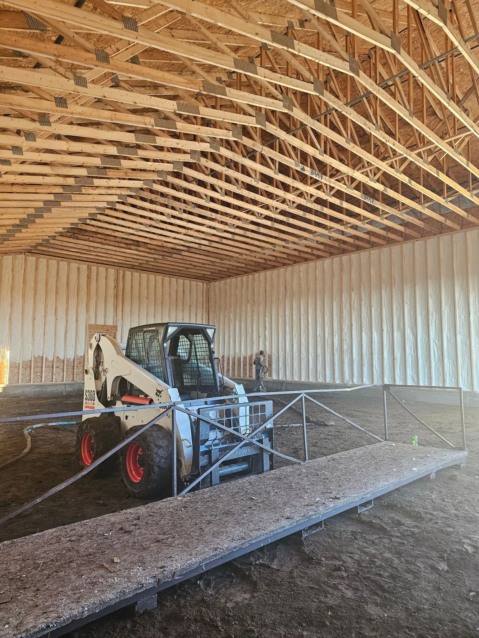 A white Bobcat skid-steer loader sits inside a large, open-framed barn with insulated walls and a gravel floor.