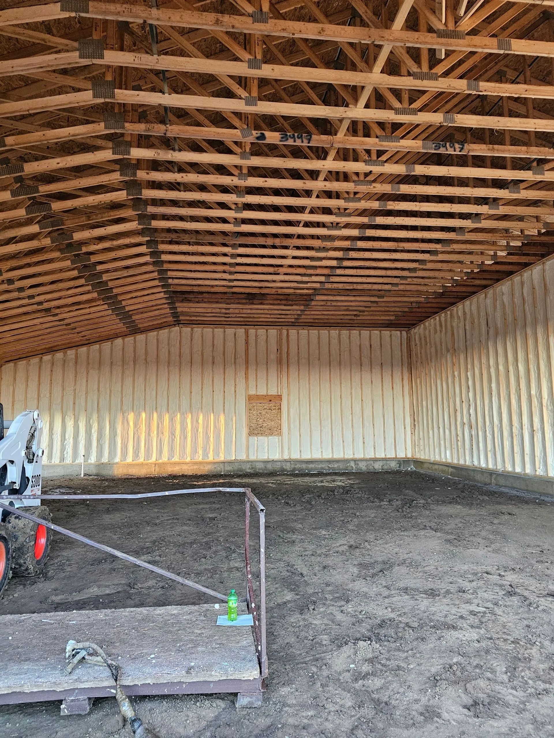 Interior of a large unfinished building with wooden rafters, spray-insulated walls, and a dirt floor.