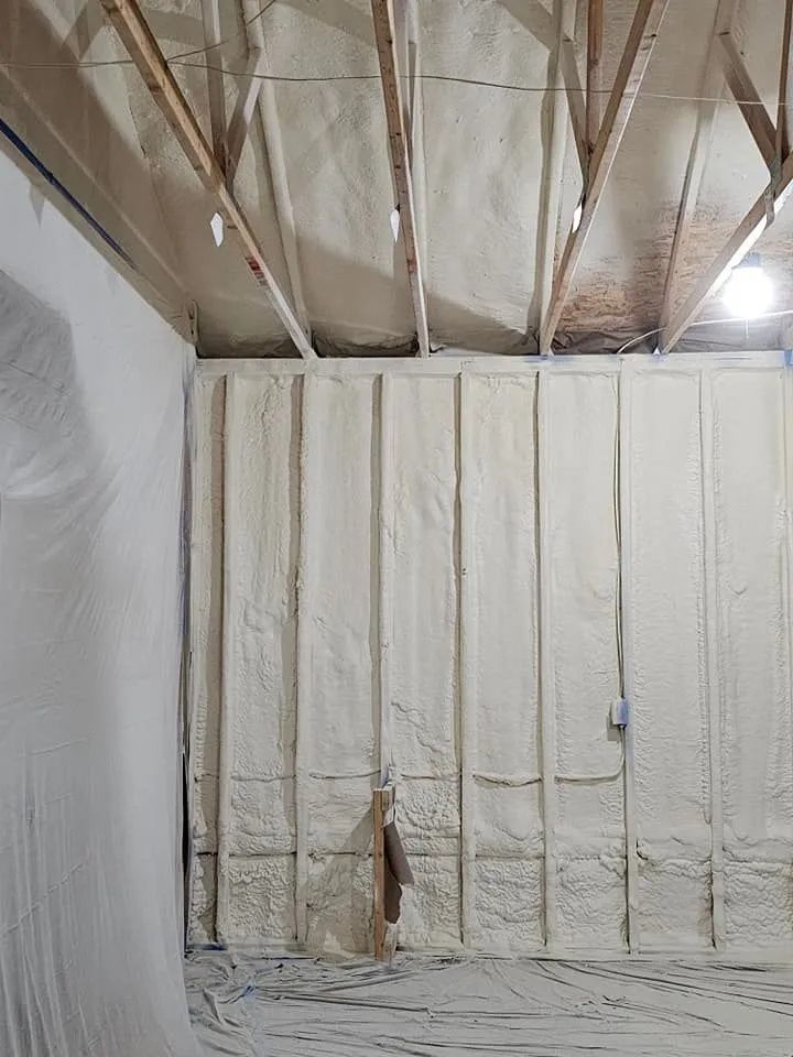 Interior view of a room under construction with spray foam insulation applied between wooden wall studs and ceiling joists.