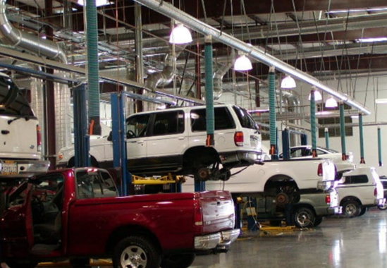 Vehicles on lifts inside a well-lit auto repair shop. A red truck is in the foreground, with white SUVs and trucks in the background.